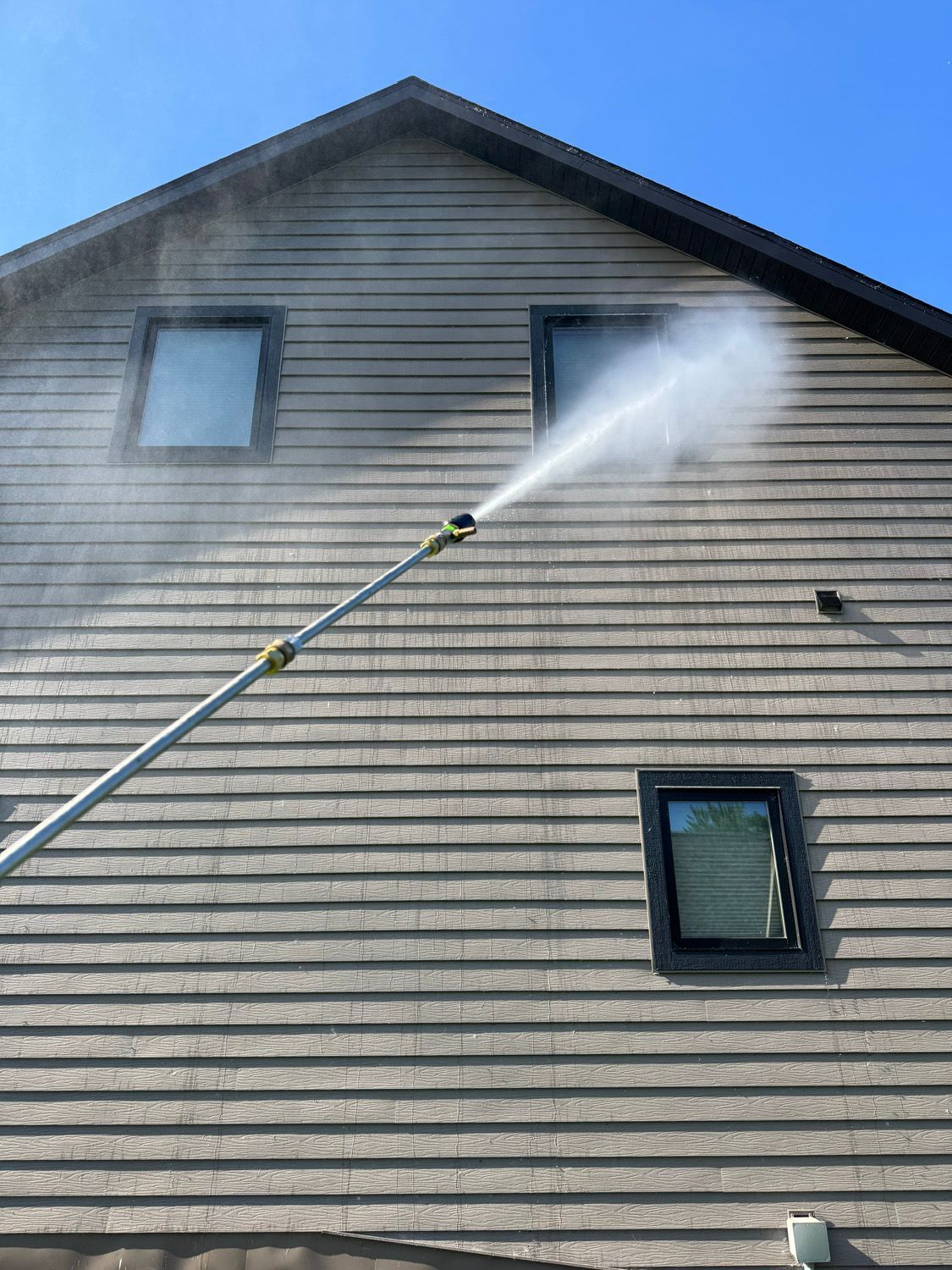 Person spraying water on tan house siding with a long pole, cleaning the exterior near the roofline.