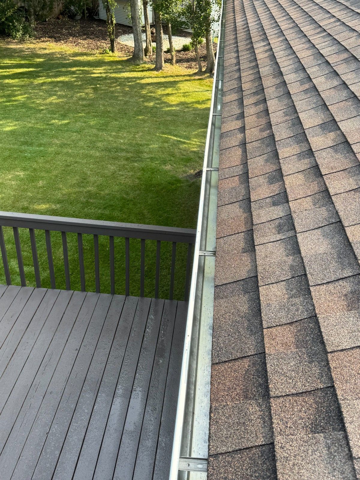 Back deck beside a sloped shingle roof, with green lawn and trees beyond the railing.