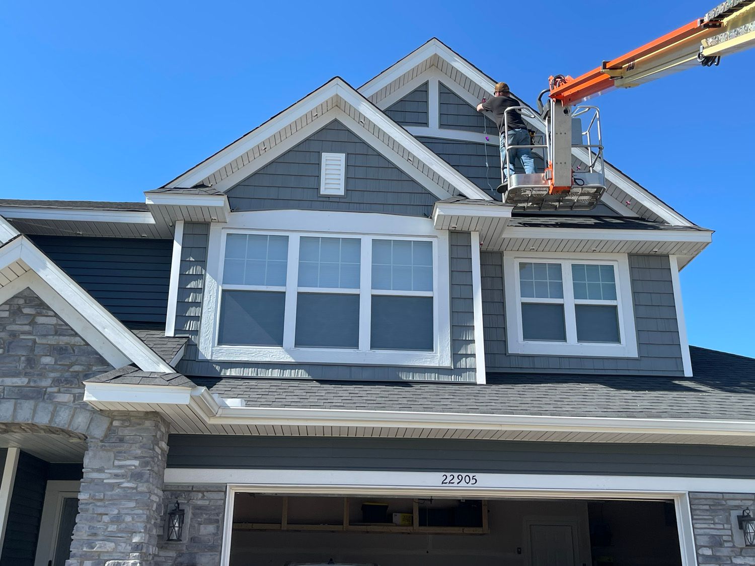 Workers on a roof using a boom lift to repair a gray house with white trim.