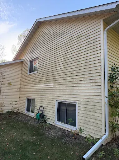 Side view of a beige house with vinyl siding, basement windows, and a white downspout beside a grassy yard.