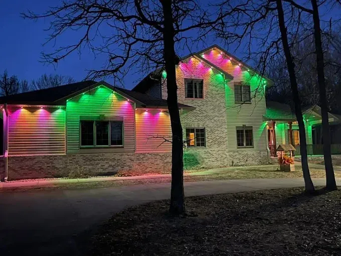 House lit with colorful Christmas lights at dusk, with trees in front and snow on the ground.