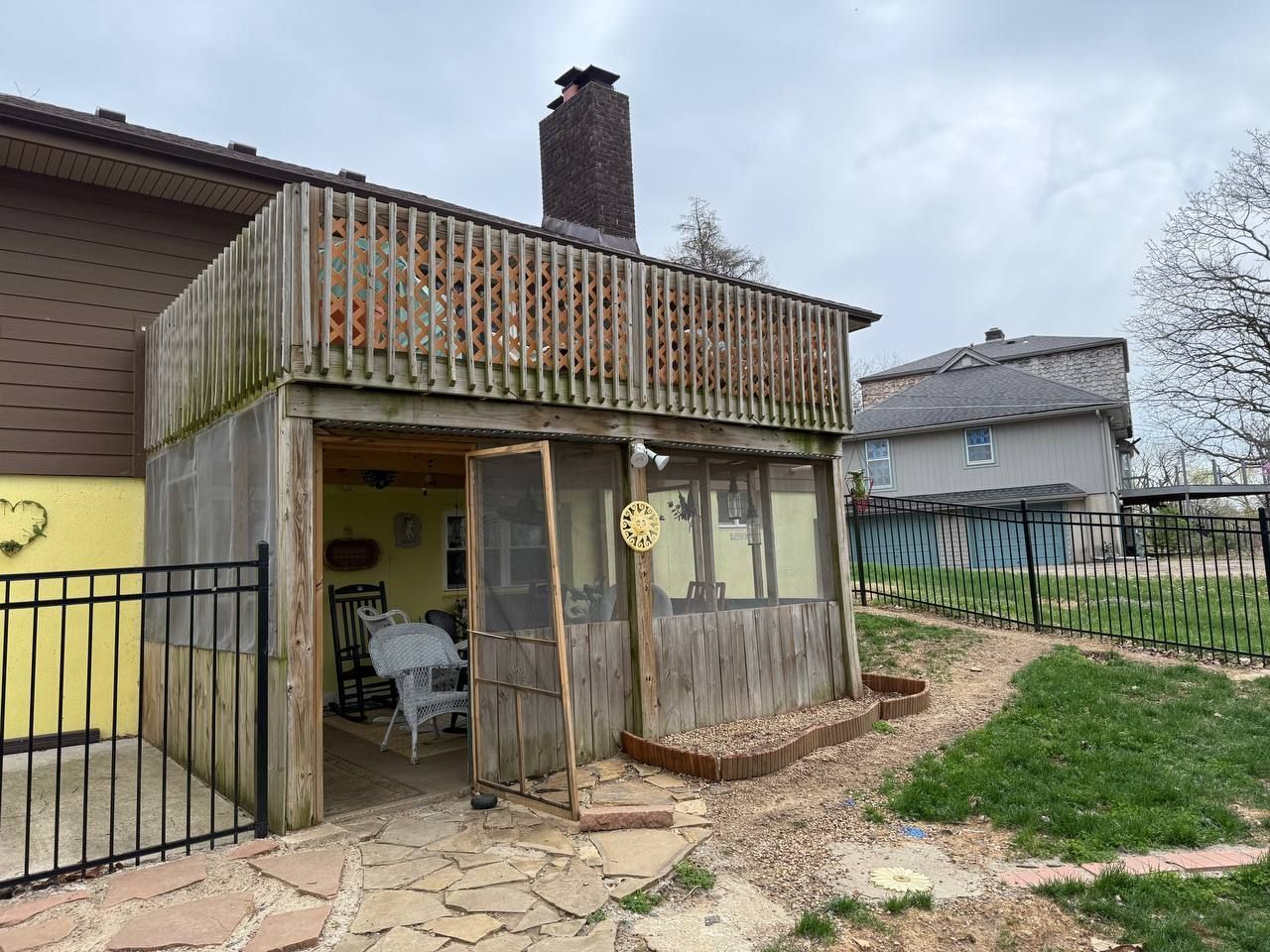 Small weathered porch attached to a house, with a chair, railing, and a chimney in a yard.