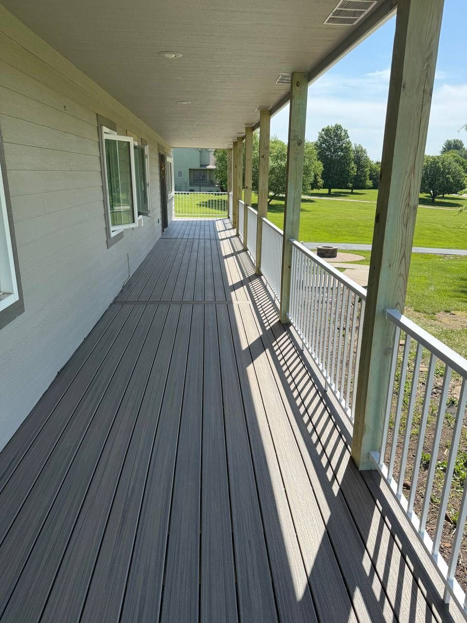 Covered apartment balcony with gray flooring, white railing, and a view of a green lawn and trees.