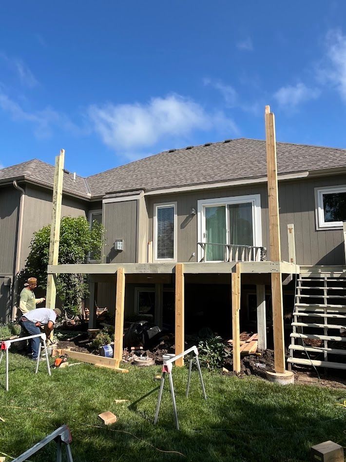 Backyard deck under construction behind a gray house, with workers and tools in the grass.