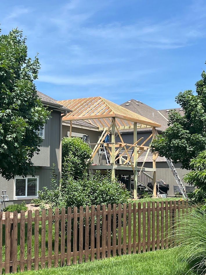 Partly built house with roof framing, surrounded by trees and a wooden fence under a blue sky