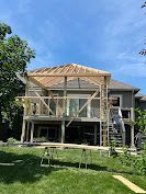 House under construction with a wooden porch and roof frame, a ladder on the right, and green grass in front.
