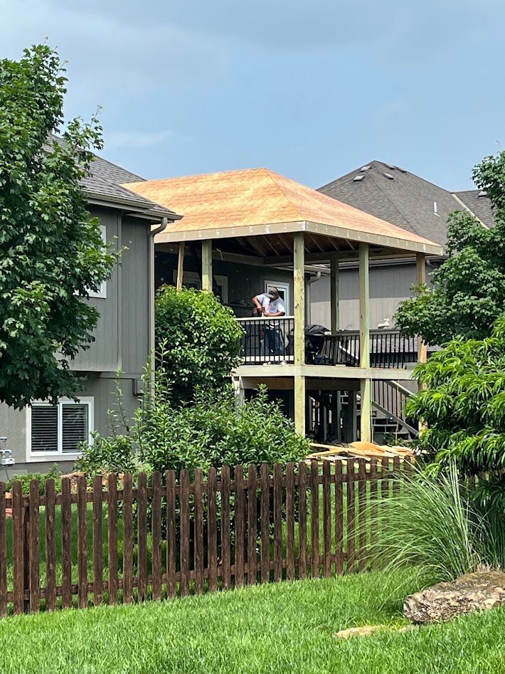 Two-story house with a covered balcony and weathered roof behind a wooden fence and green lawn