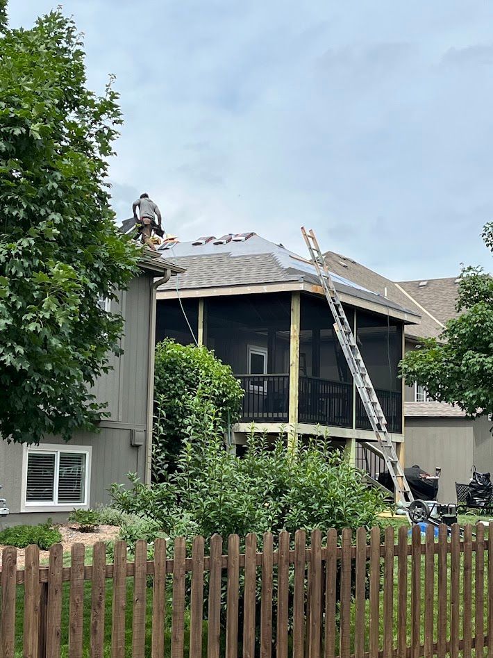 Workers repairing roof on a two-story house, with ladders and shingles visible.