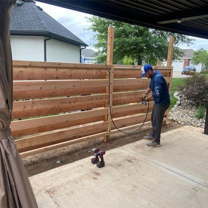 Person watering a newly built wooden fence beside a driveway, with a small dog nearby