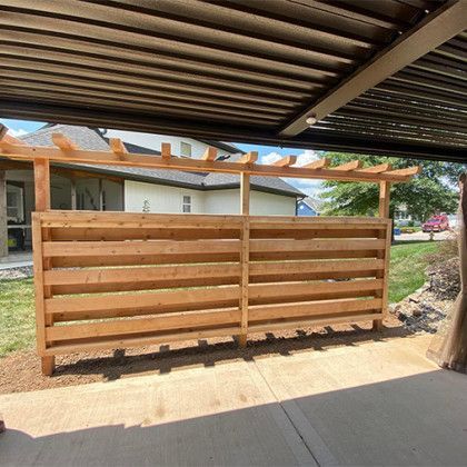 Wooden slat fence under a carport, with a house and yard in the background.