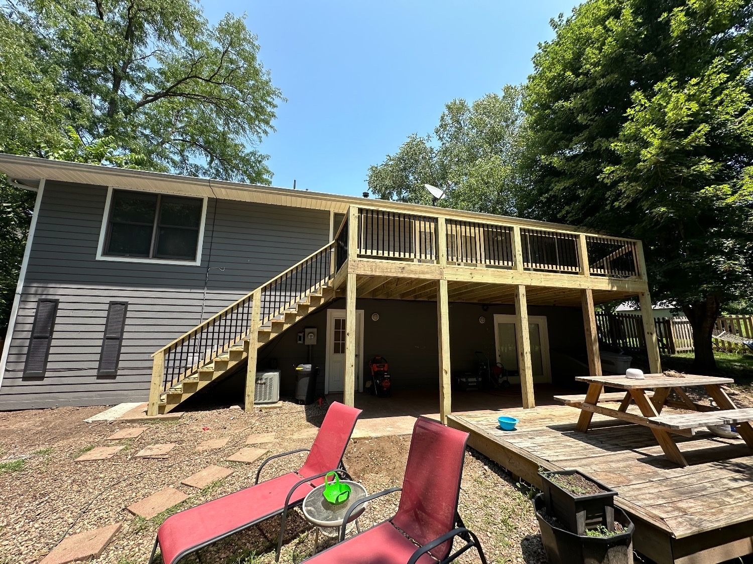 Back deck under construction beside gray house, with stairs, picnic table, and lawn chairs in a yard