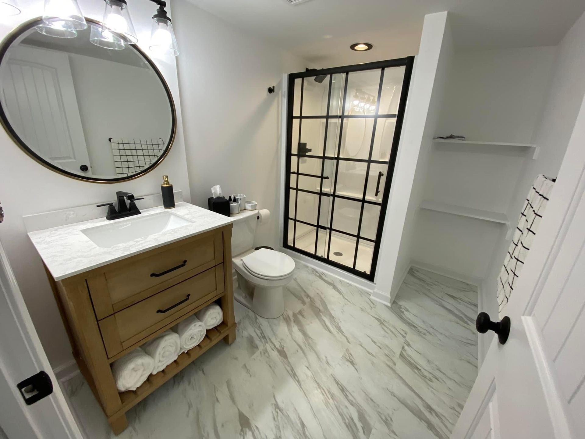Modern bathroom with wood vanity, round mirror, black-framed shower door, and marble-look floor tiles