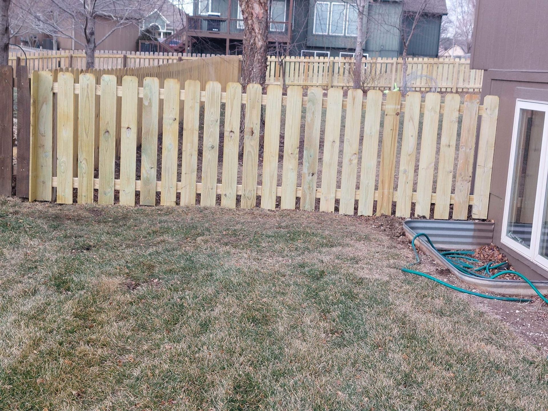 Backyard with a light wood fence, patchy grass, and a coiled hose near a house window
