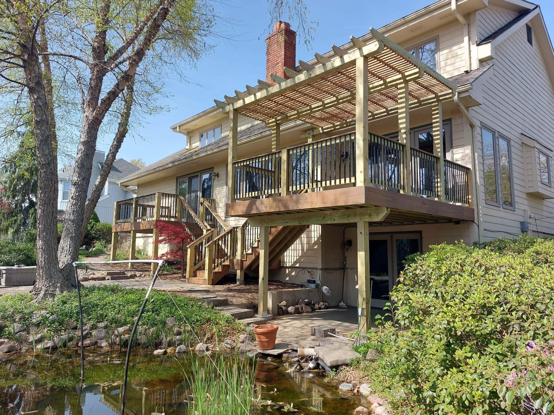 Brick house with a covered deck and pergola beside a small pond, surrounded by trees and shrubs.