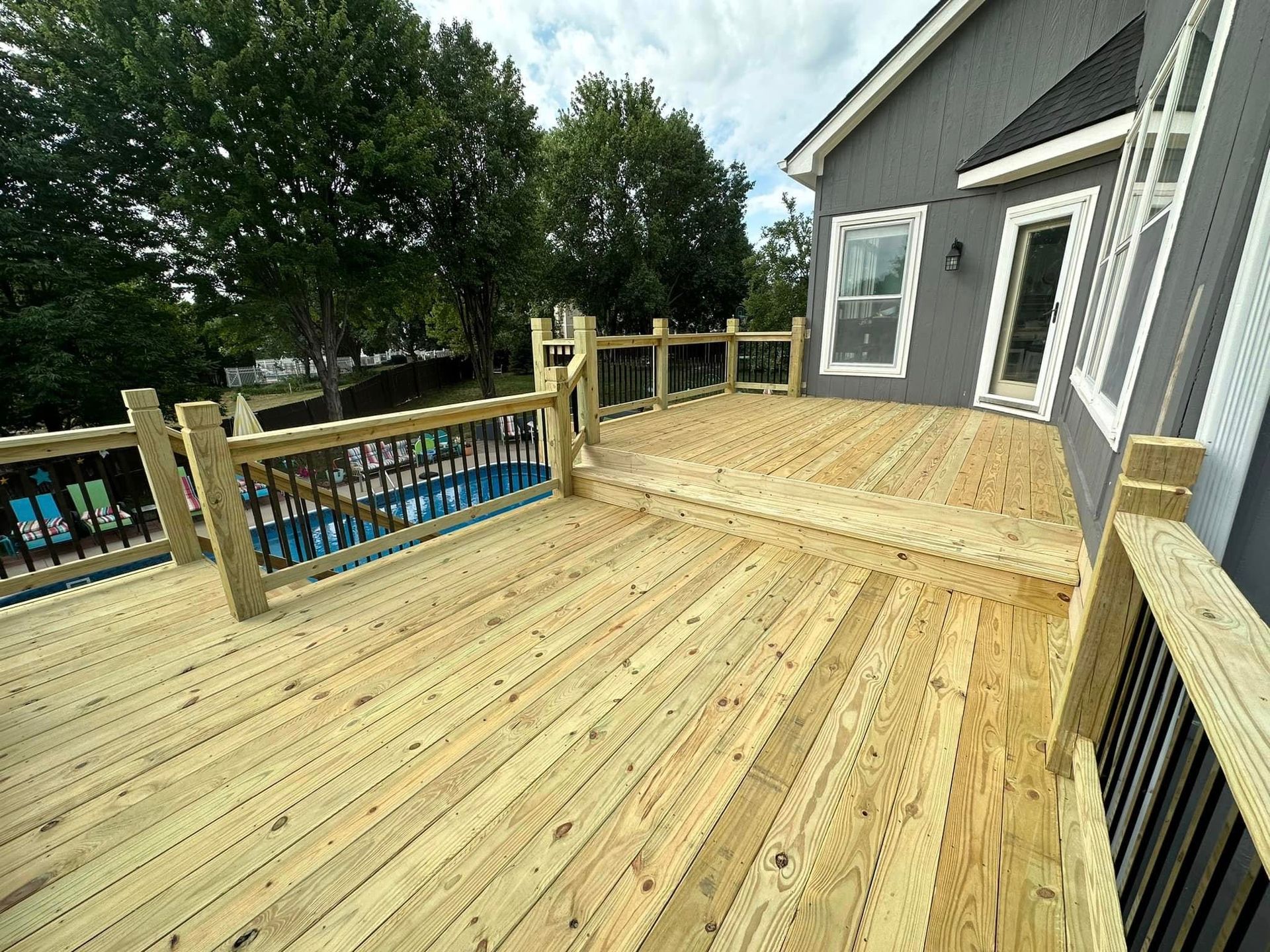 Wooden deck with railings beside a house, overlooking a backyard with a pool on a sunny day