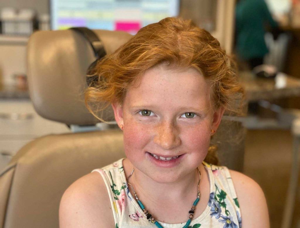 Girl with red hair smiles in a dentist's chair, wearing a floral top, dental office setting.