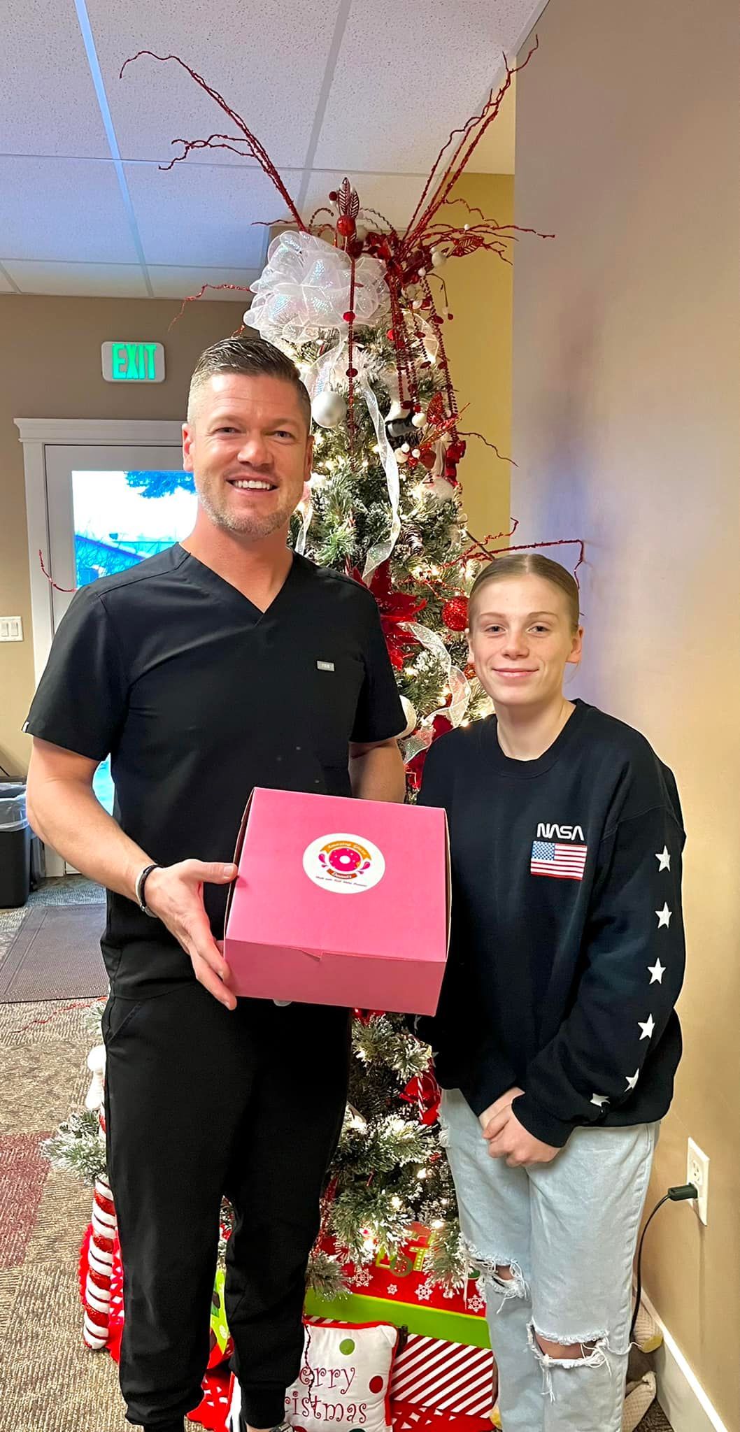 Man in black scrubs and young person by a Christmas tree, holding a pink gift box; smiling.