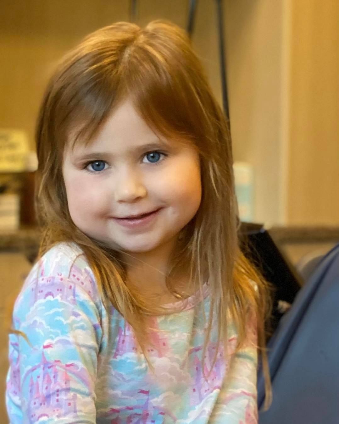 Girl with long, red hair and blue eyes smiles; wears a colorful shirt.