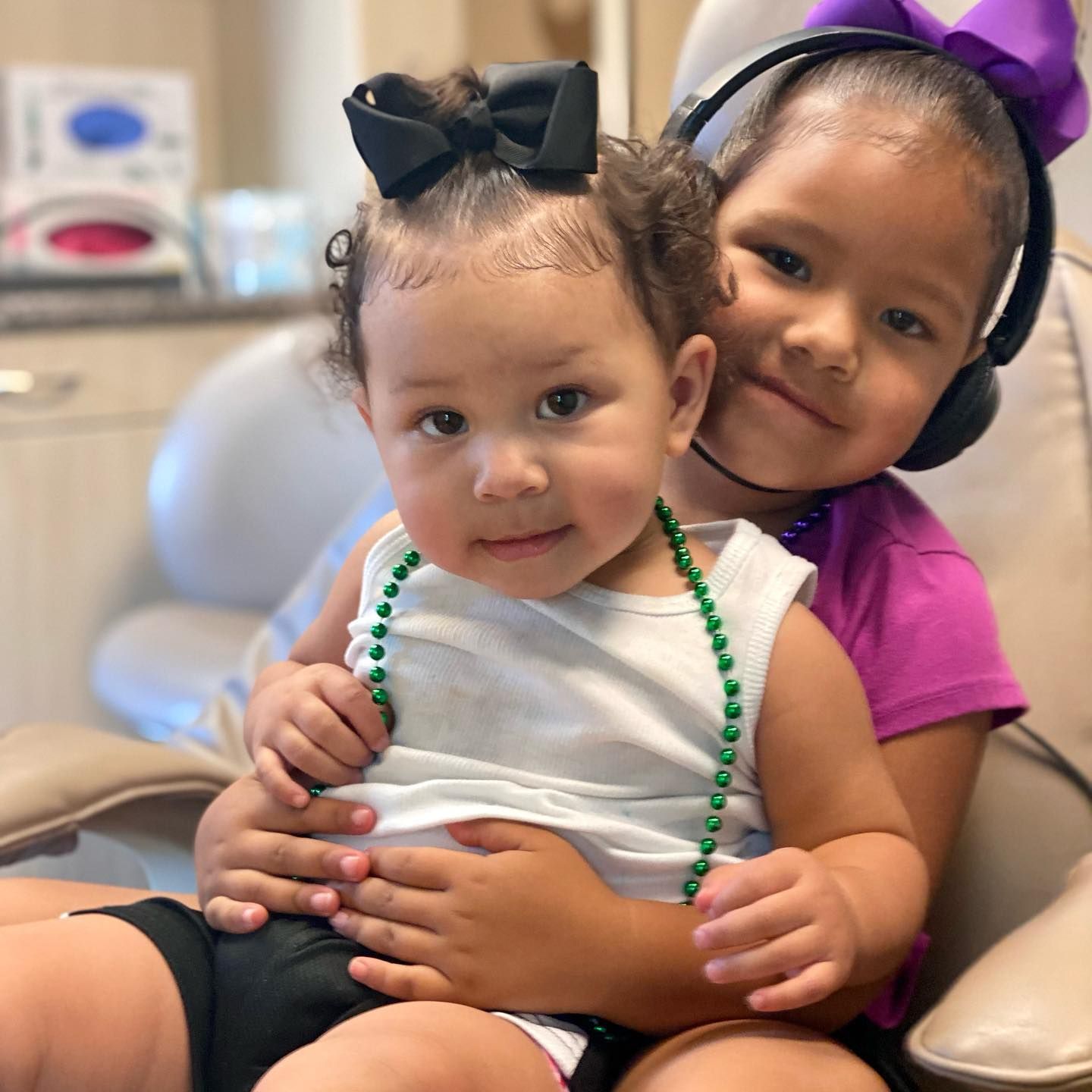Two children sit together, smiling. One wears a bow and holds a bead necklace, the other wears a headset and a bow.
