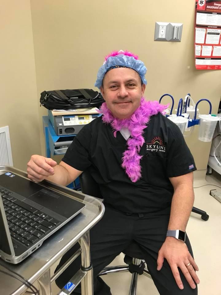 Man in scrubs with a pink boa and shower cap, sitting at a desk with a laptop.