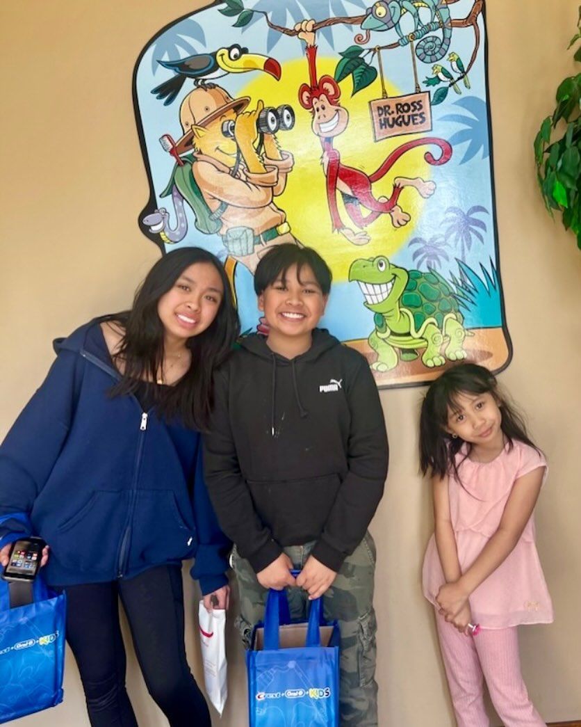 Three children pose with gift bags in front of a jungle-themed mural, smiling.