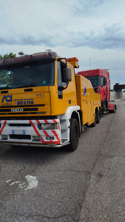 Un carro attrezzi solleva la cabina bianca di un semirimorchio sulla corsia di emergenza di un'autostrada, con un operaio nelle vicinanze.