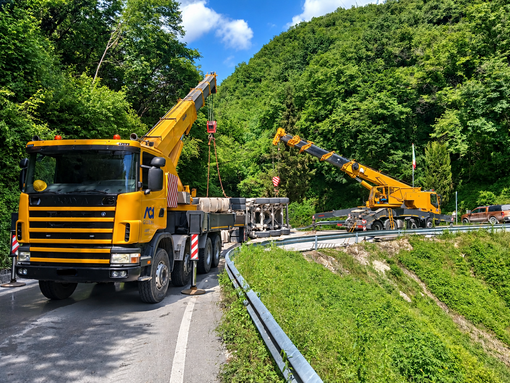Operaio inginocchiato accanto alla ruota di un camion durante una riparazione stradale, con un furgone giallo e un cono stradale nelle vicinanze.