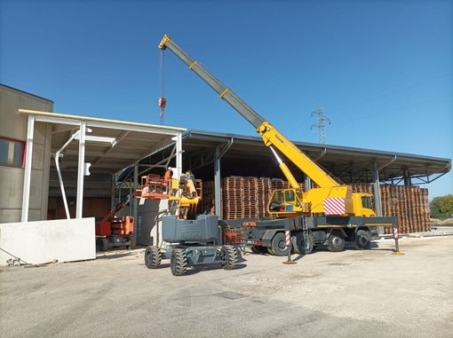 Una gru da costruzione solleva un blocco di cemento mentre un operaio con un giubbotto arancione guida il carico in cantiere.
