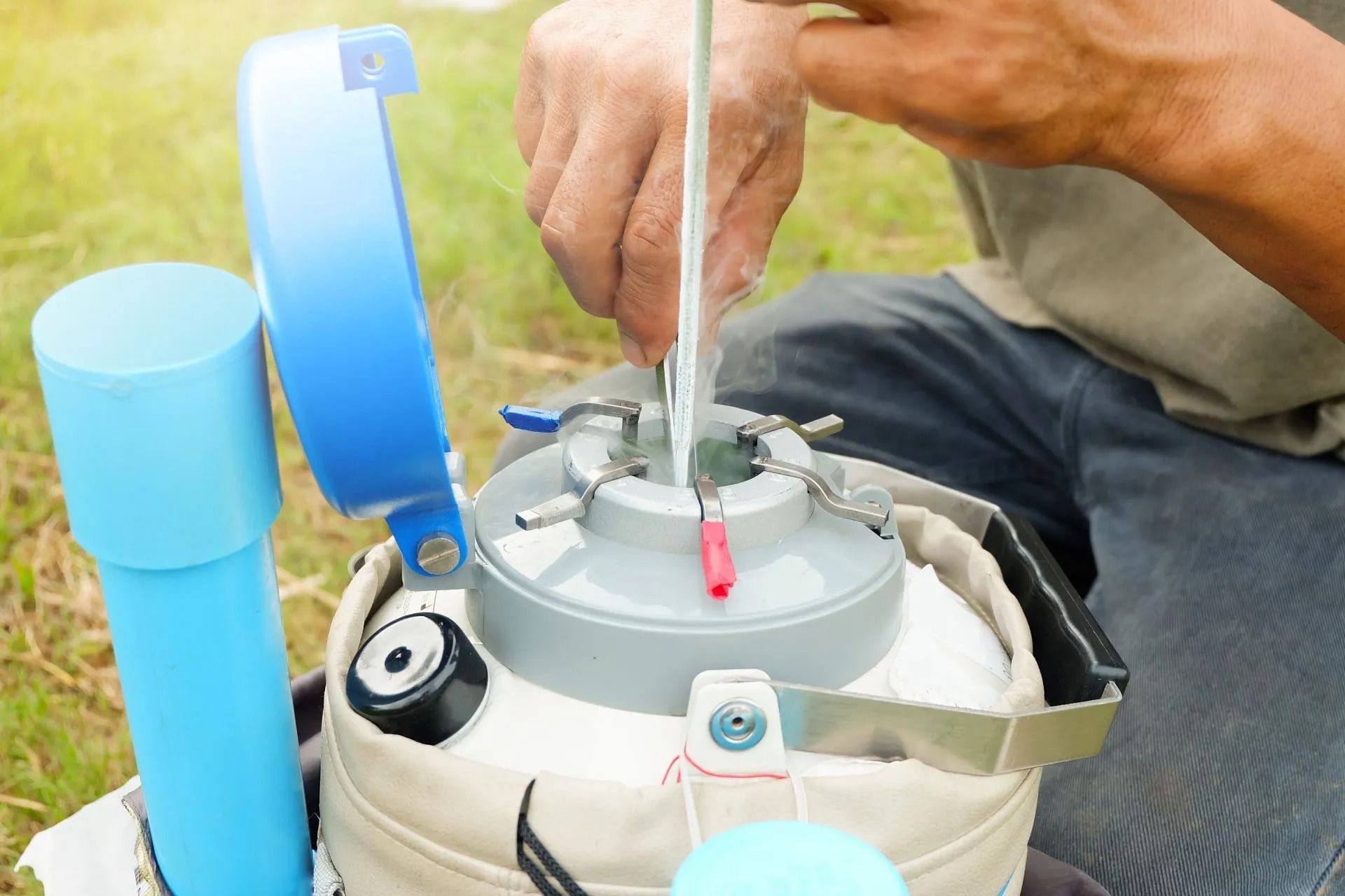 Hands adjusting a gray camping kettle with blue cups beside it on grass
