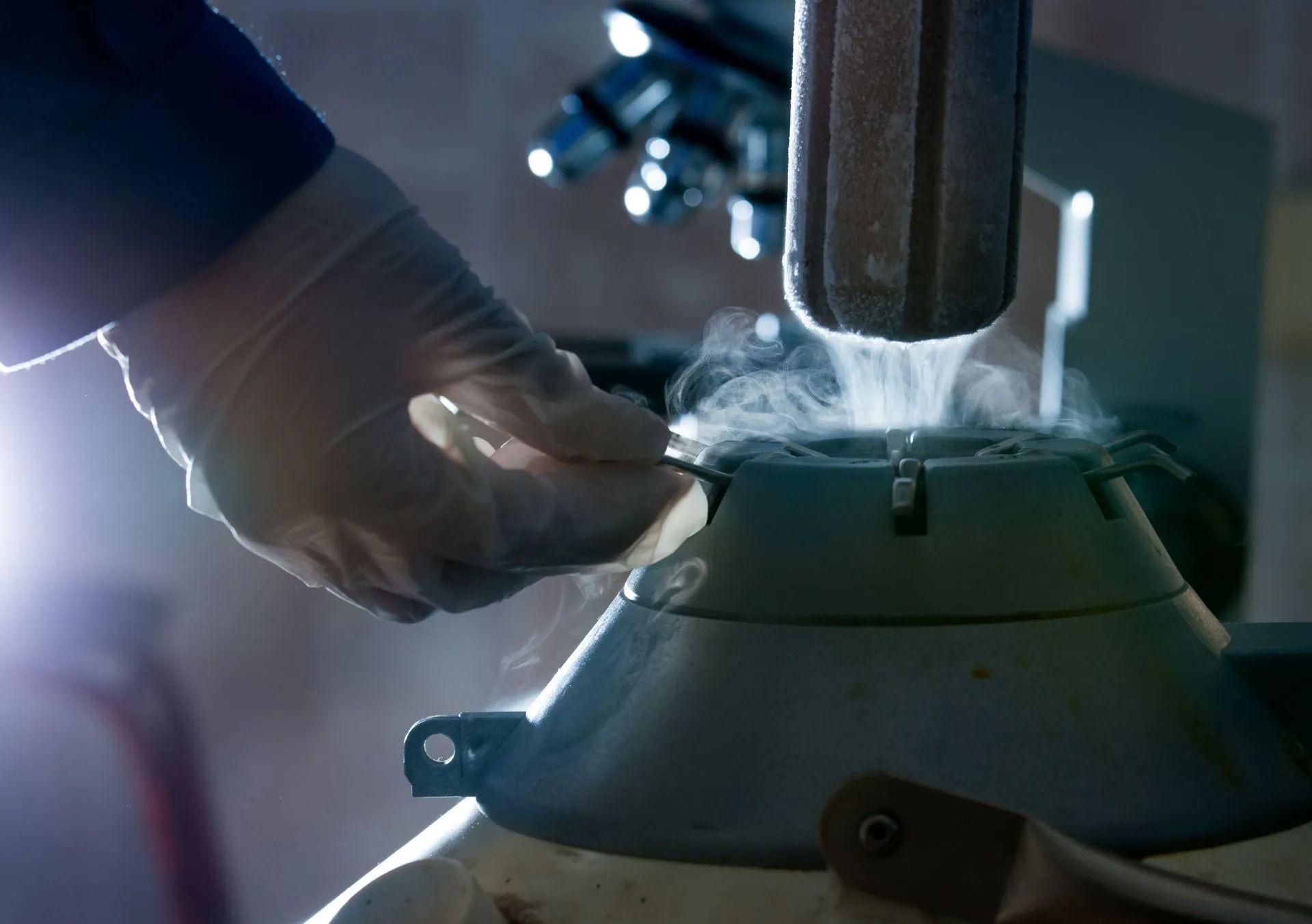 Gloved hand pouring liquid into a metal machine as steam rises in a workshop
