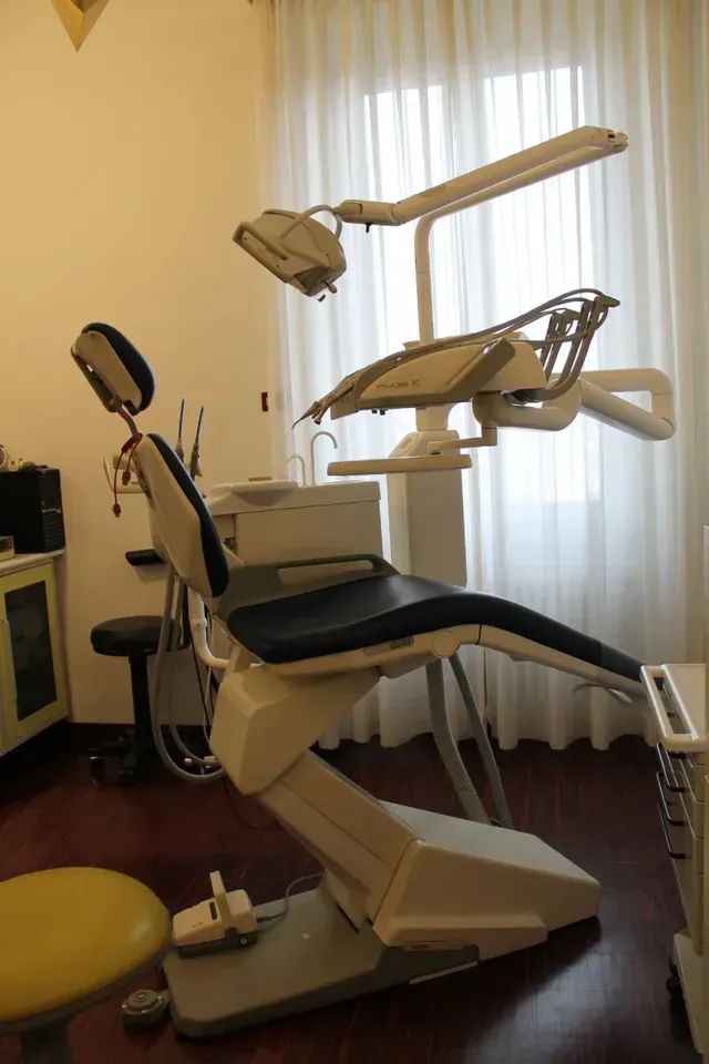 Poltrona odontoiatrica e lampada da visita a soffitto in una piccola stanza clinica con finestra coperta da una tenda.