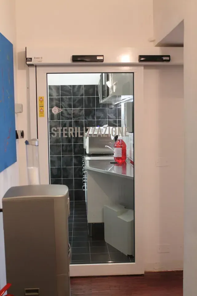 Ingresso di una stanza di sterilizzazione con porta a vetri, piastrelle nere e un dispenser di sapone rosso all'interno.