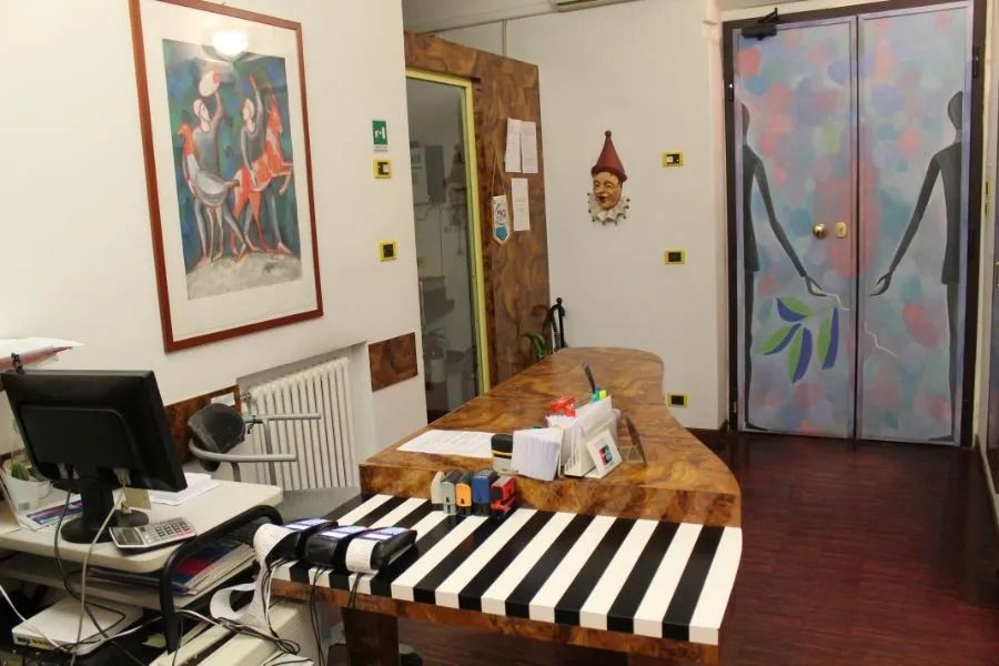 Interno di un ufficio con scrivania, computer, tovaglia a righe, quadri alle pareti e tende colorate.