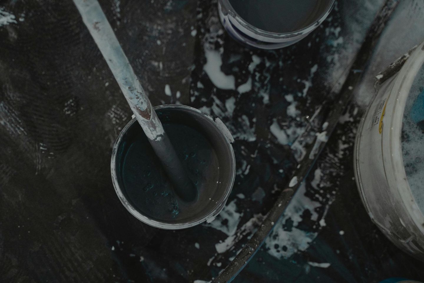 Paint can with dark blue paint, stirrer, and other open cans on a dark surface.
