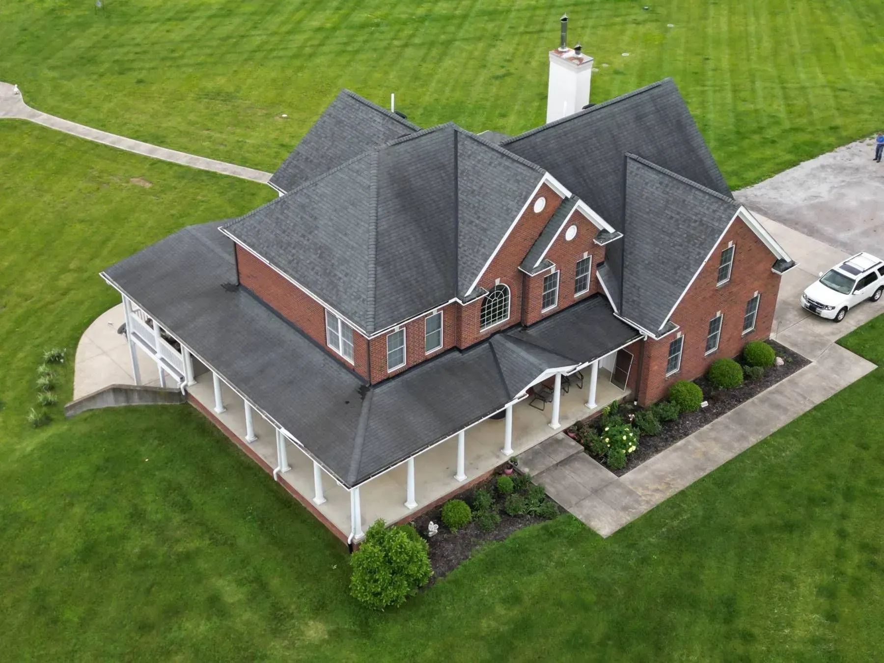 Aerial view of a brick house with a dark gray roof and wraparound porch on a green lawn.