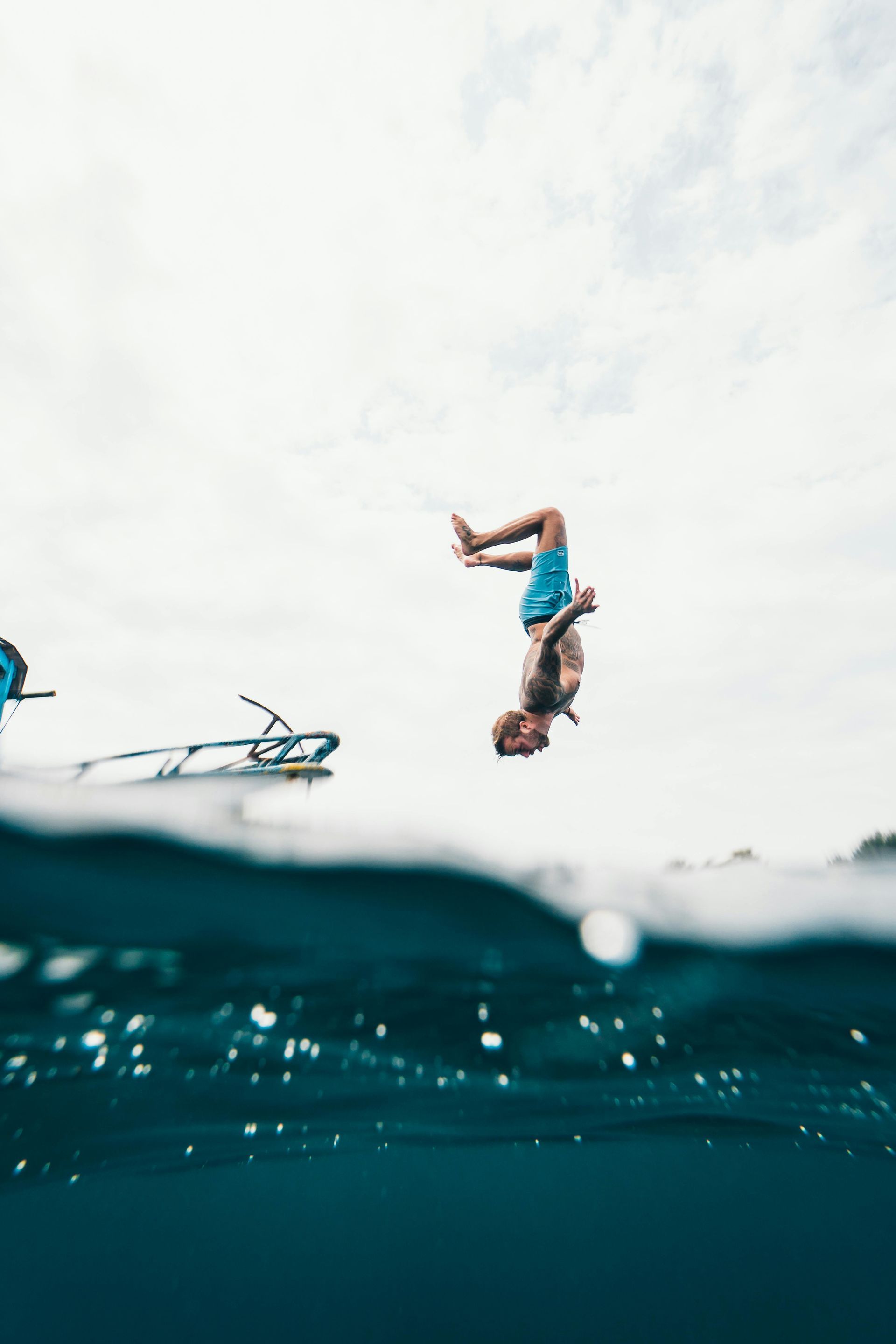 A person doing a backflip into blue water with a partially visible bicycle. Overcast sky.