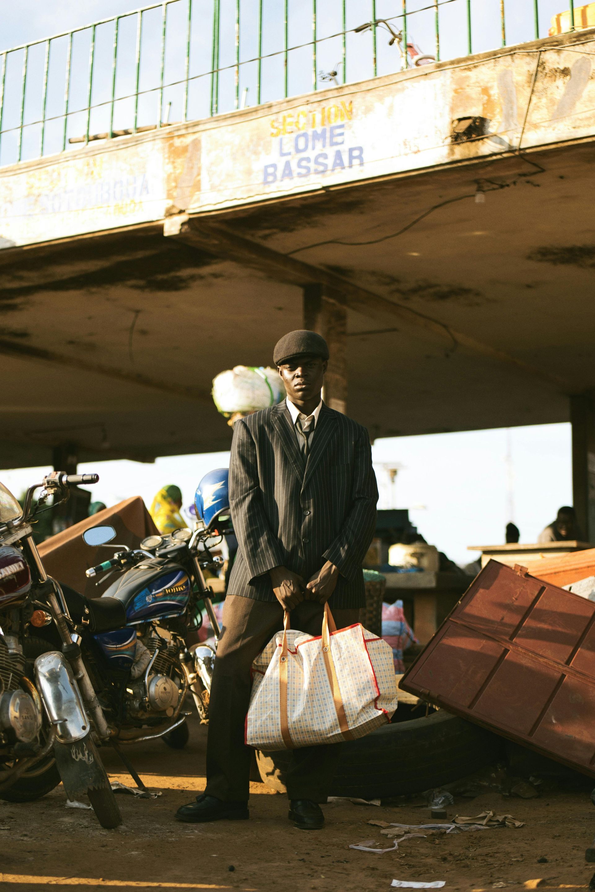 Man in jacket and cap holds a bag, standing by motorcycles and debris under a concrete structure. 