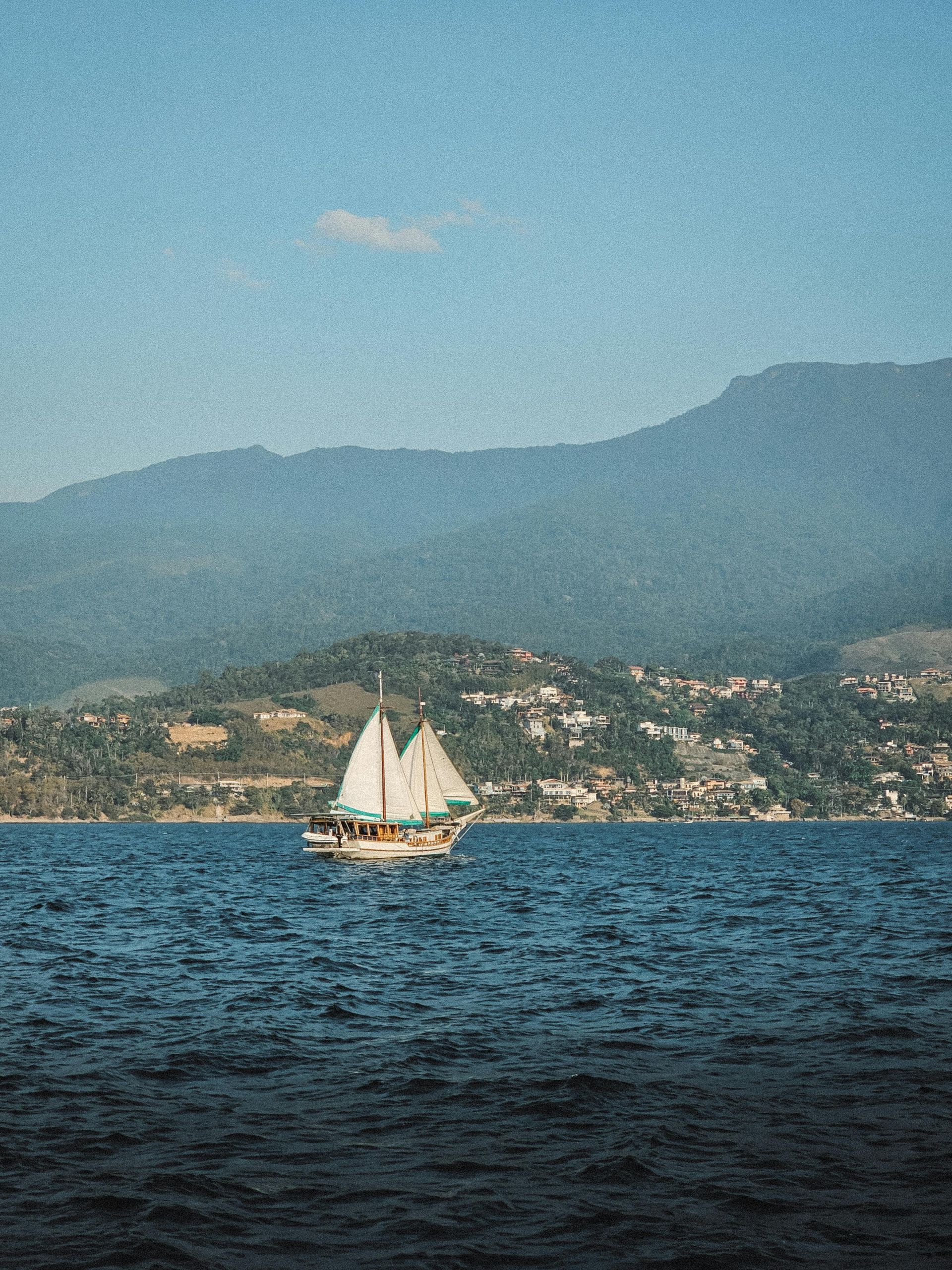 Sailboat on blue water with a mountain backdrop and a town along the shore.