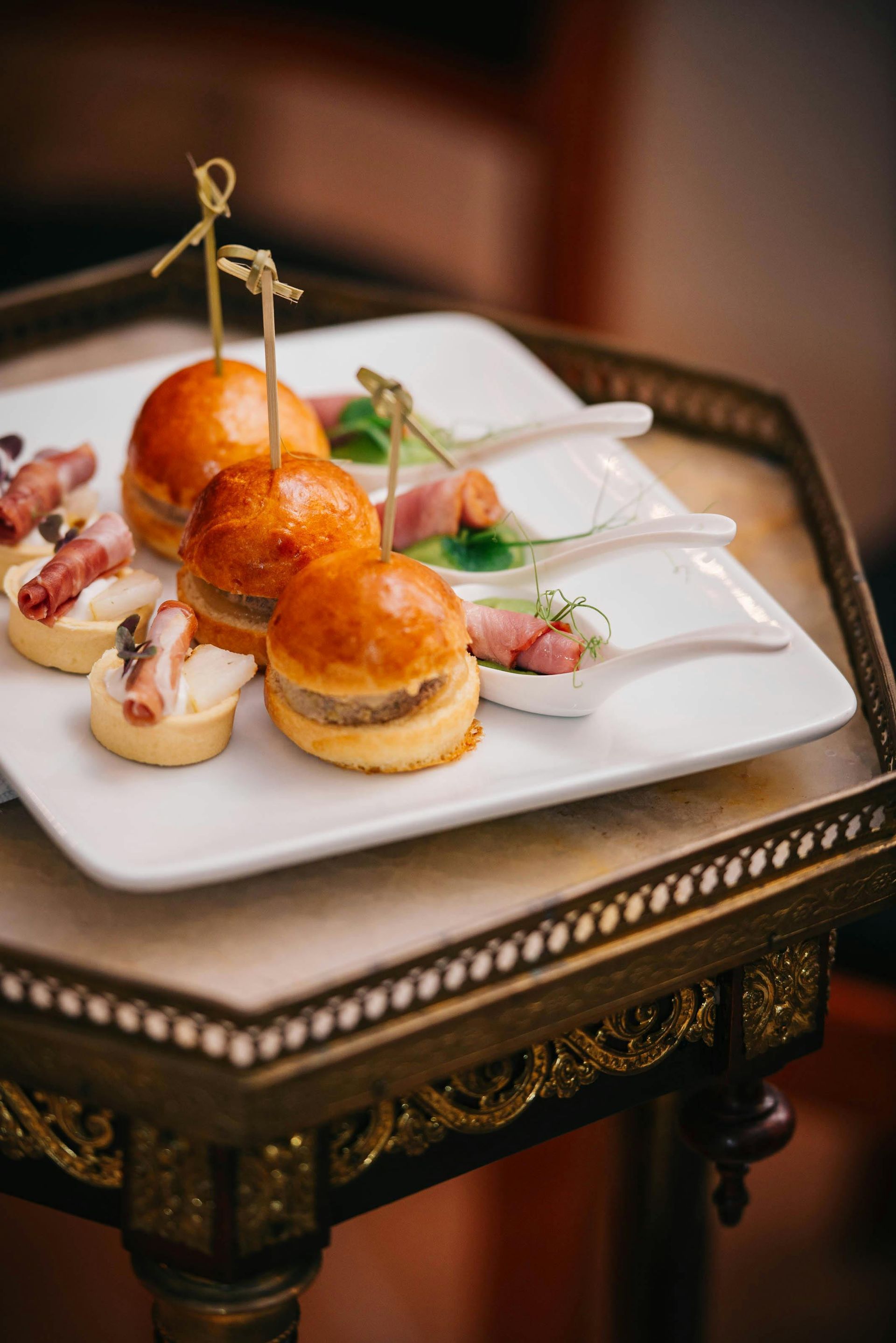 Appetizers on a white plate atop a decorative tray: mini burgers, tarts, and prosciutto bites.