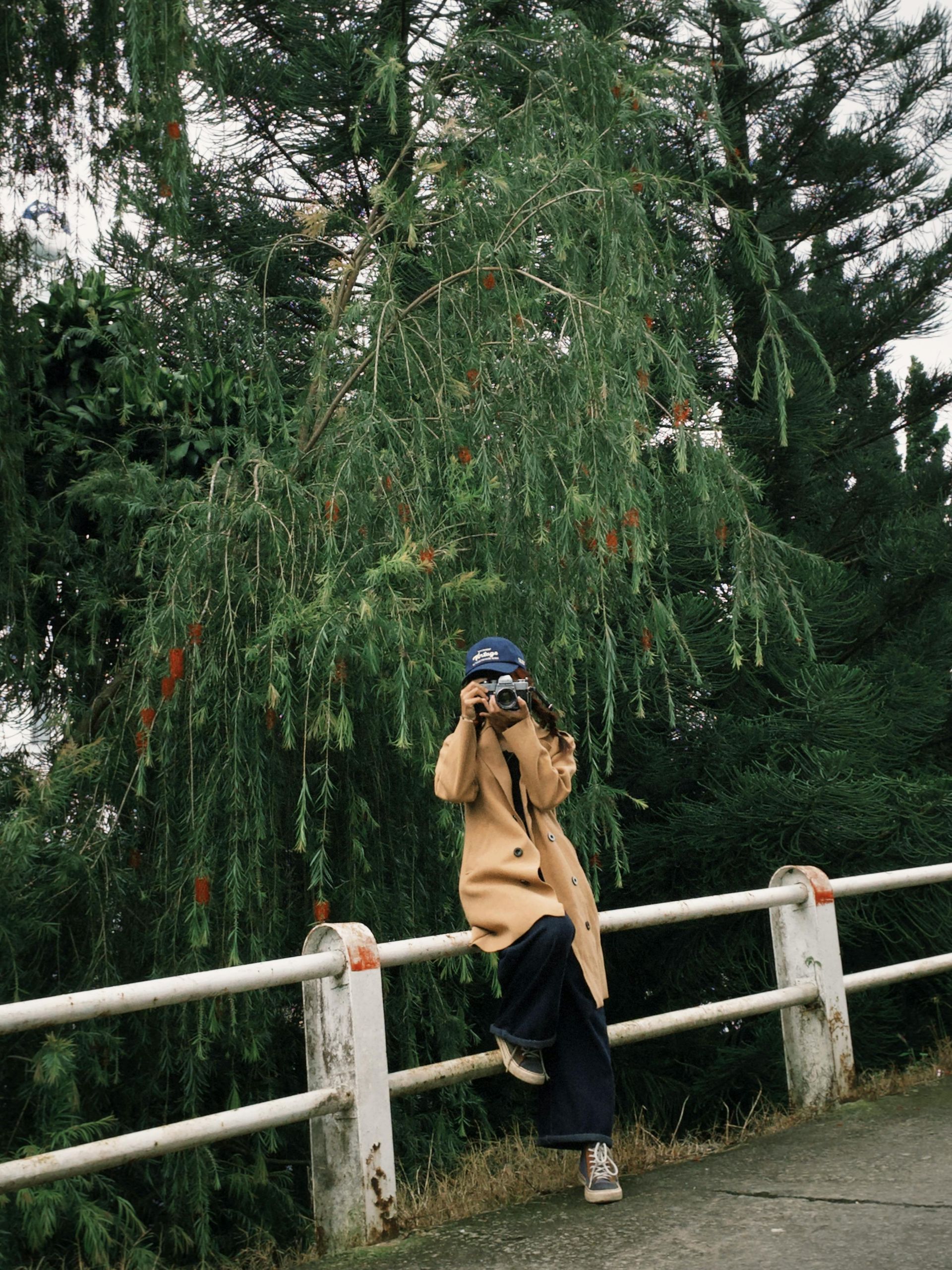 Person in tan coat and blue hat sitting on a white railing, holding a camera, with trees in the background.