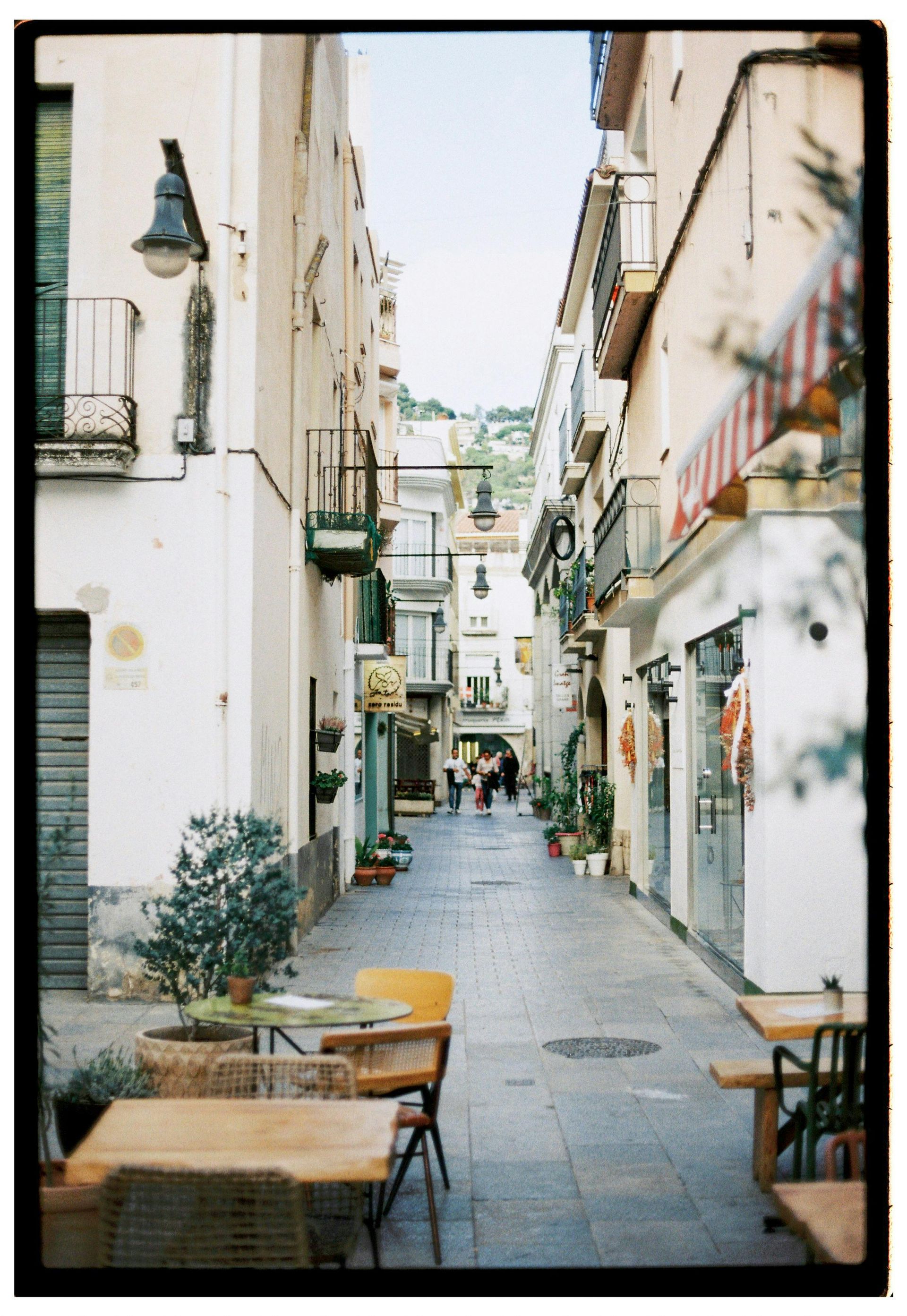 Narrow European street with buildings, outdoor seating, and people walking.