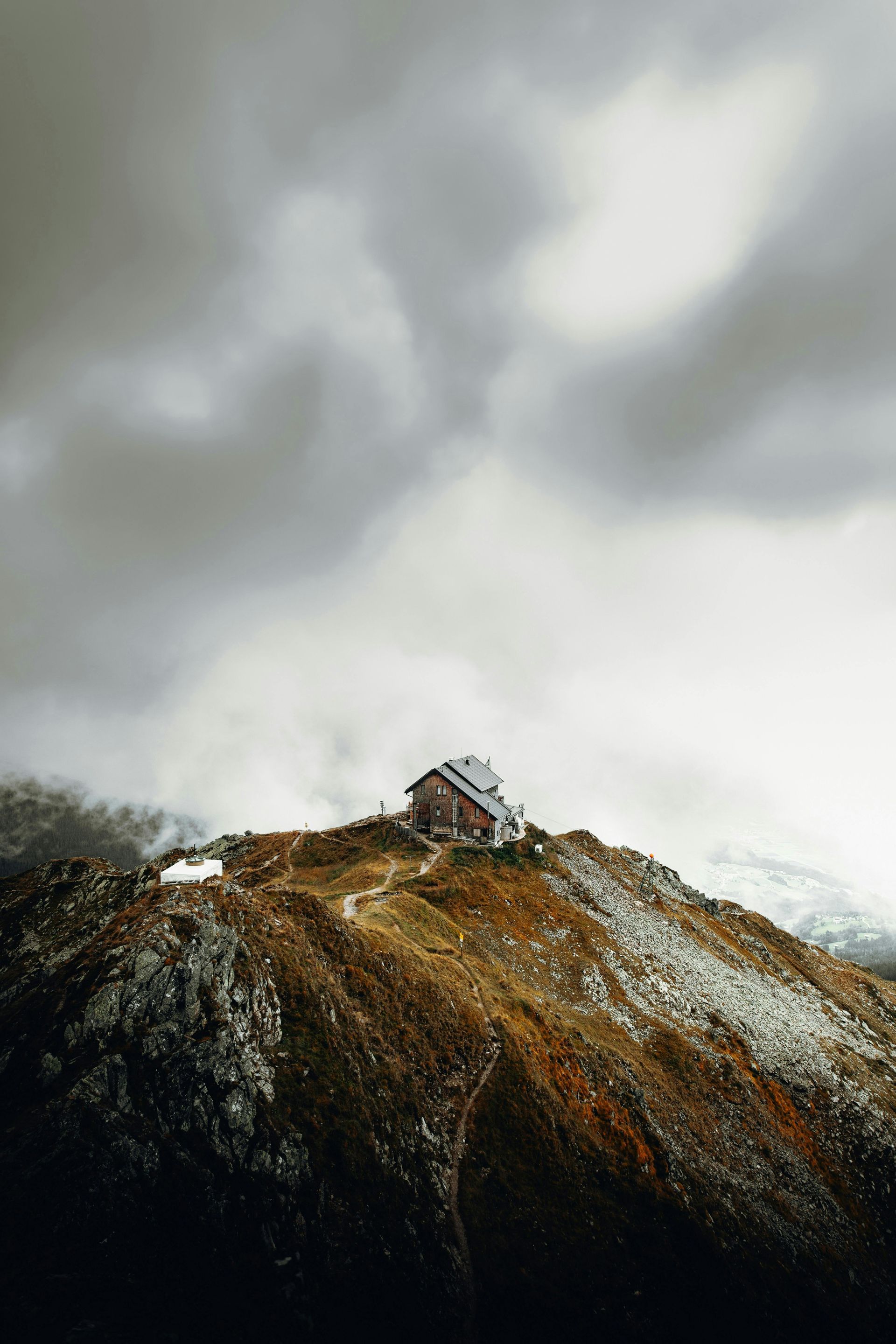 Wooden cabin atop a mountain, under cloudy sky. Snowy patches, brown and gray terrain.