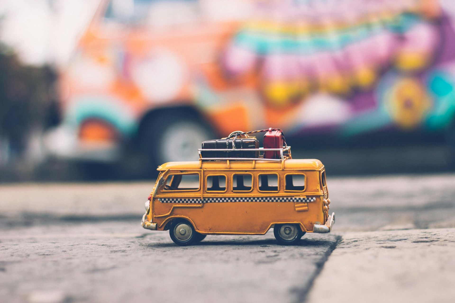 Yellow toy bus with luggage on the roof, on a street. Colorful painted bus in background.