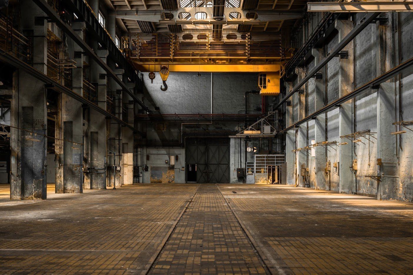 Empty industrial building interior; overhead crane, brick floor, aged concrete columns.