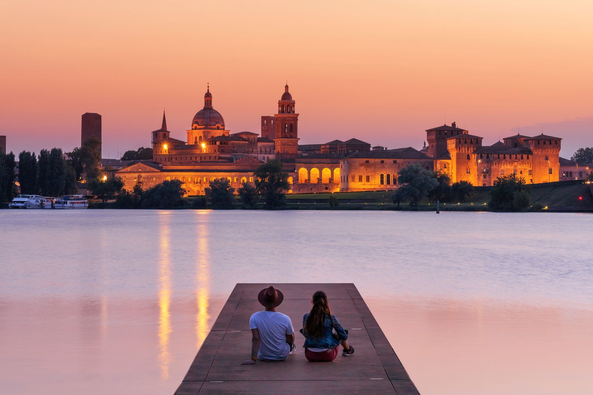 Two people are sitting on a jetty overlooking a lake at sunset.