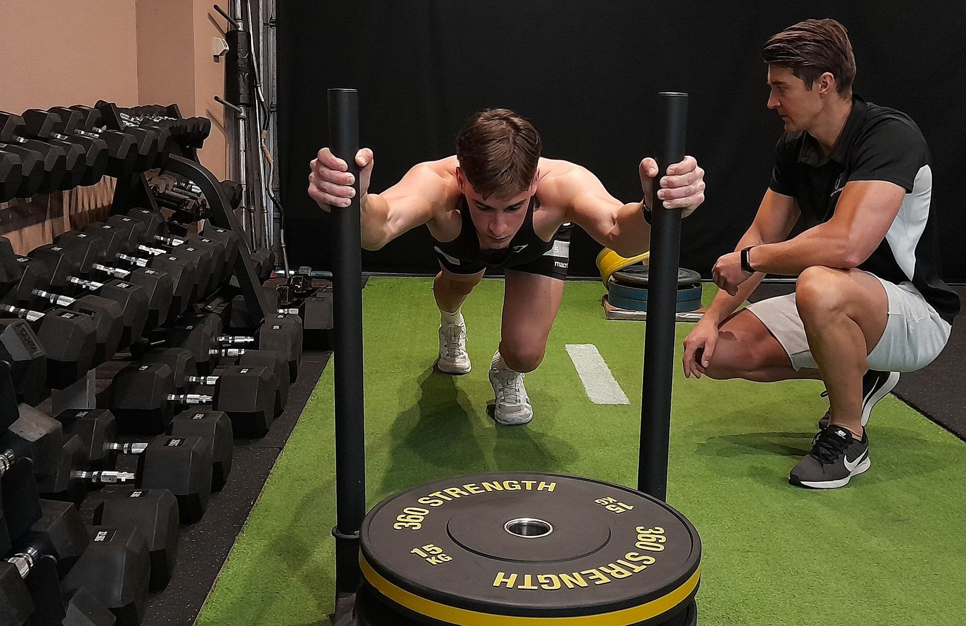 Man pushing weighted sled in a gym, assisted by a coach.