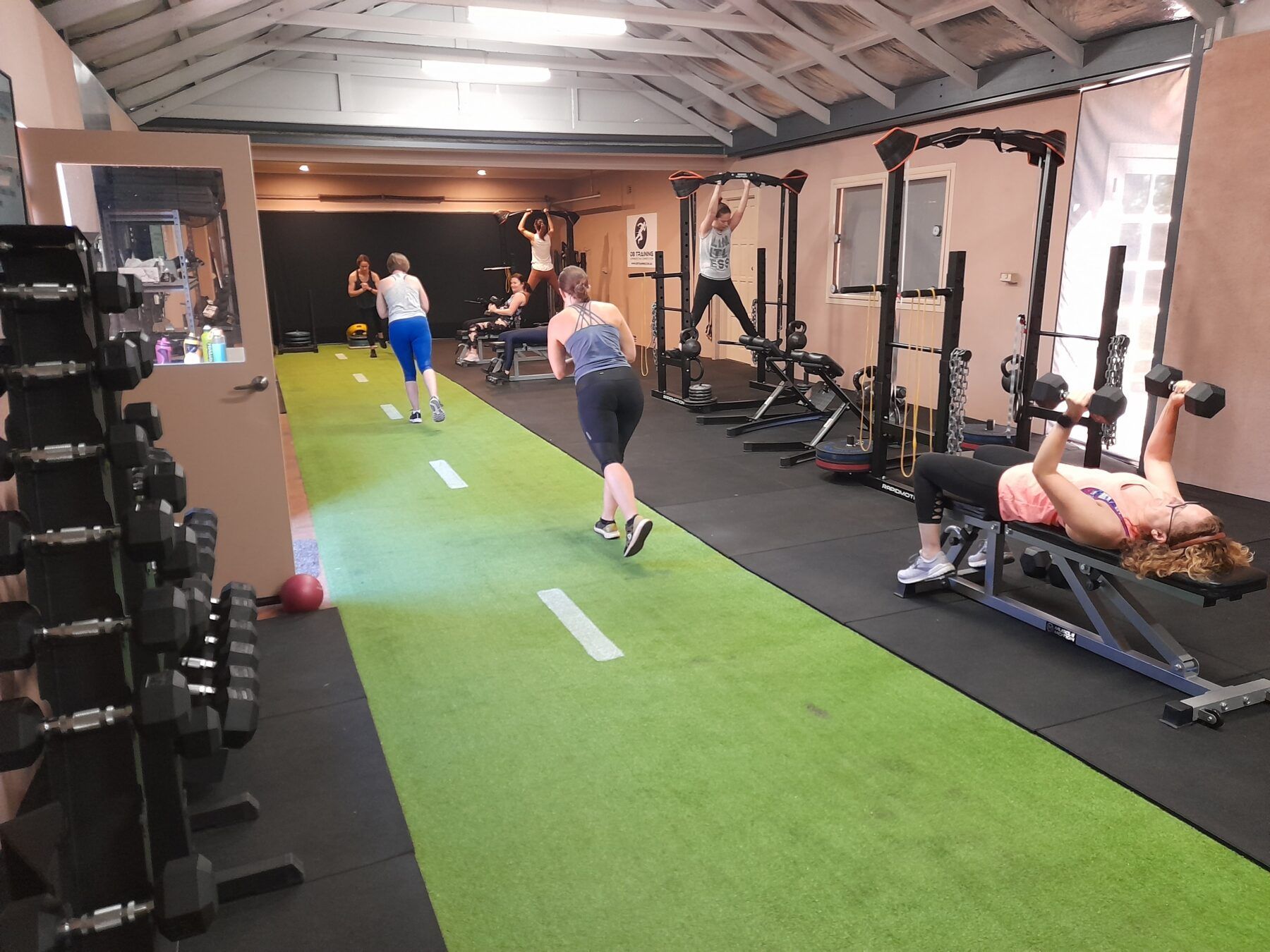 People exercising in a gym with a green turf lane, weights, and workout machines.