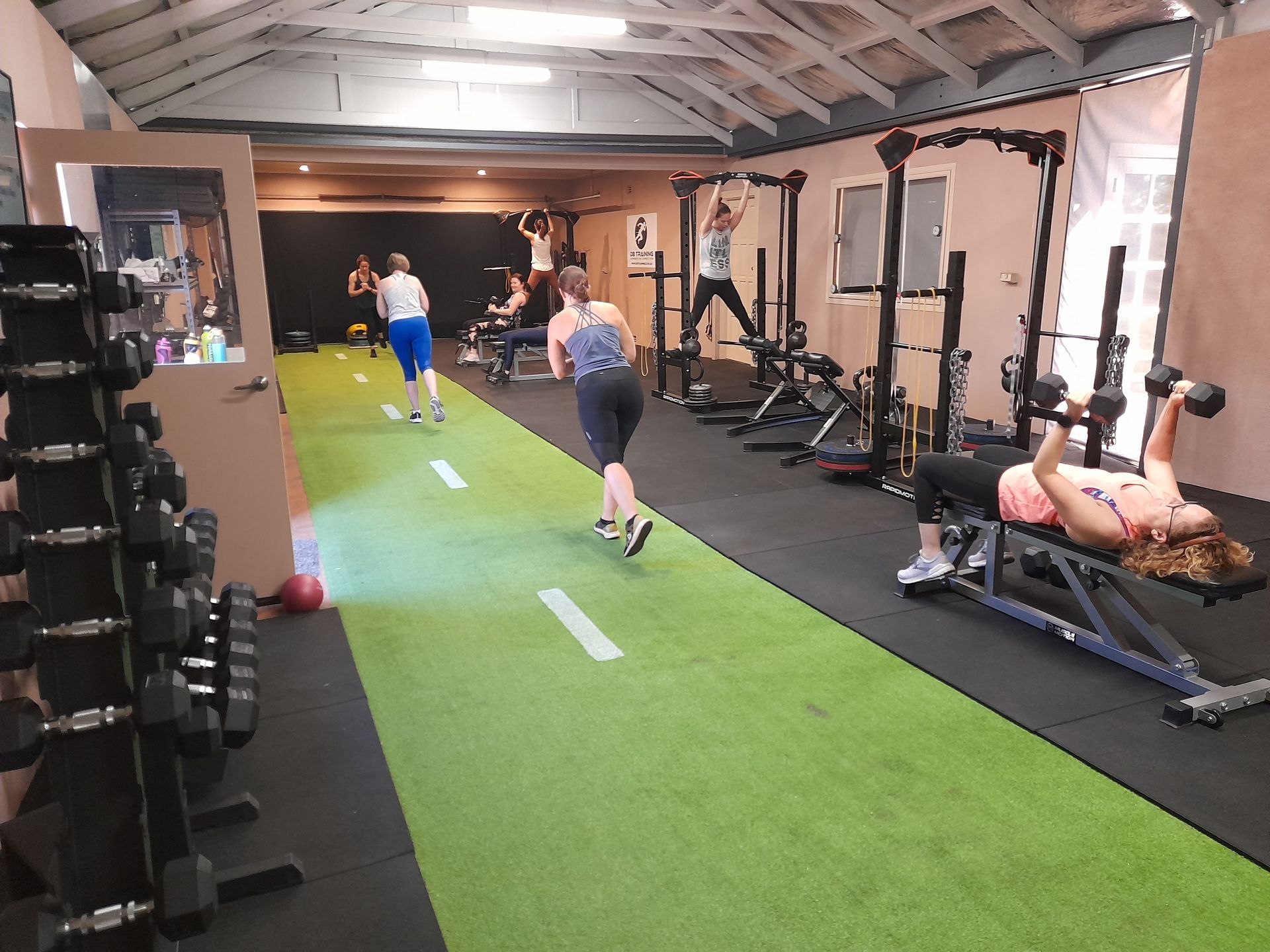 Indoor gym with a green turf lane, people exercising, dumbbells, and workout benches.
