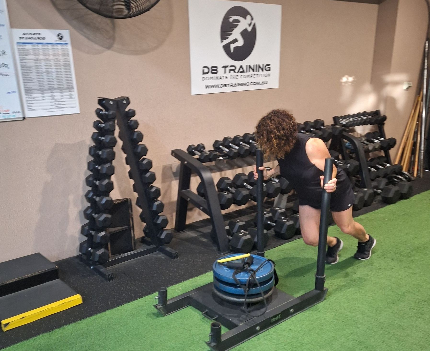 Person training in a gym, pushing a weighted sled on green turf beside dumbbells and a DB Training sign
