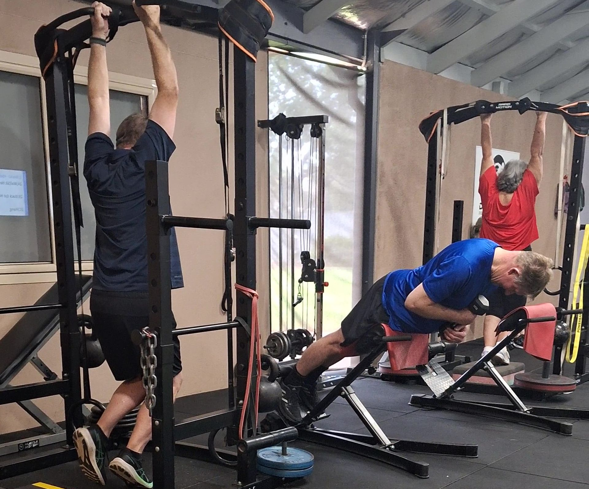 People exercising in a gym: one hangs from a pull-up bar, others use a decline bench and cable machine.